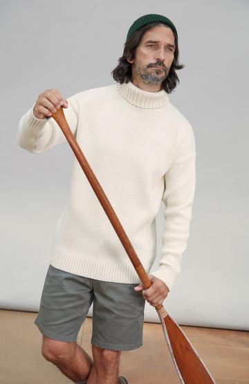 A man wearing a cream wool submariner ribbed turtleneck sweater, gray shorts, and a dark green beanie, holding a wooden paddle against a light gray studio background. Made in England by Paul James Knitwear.