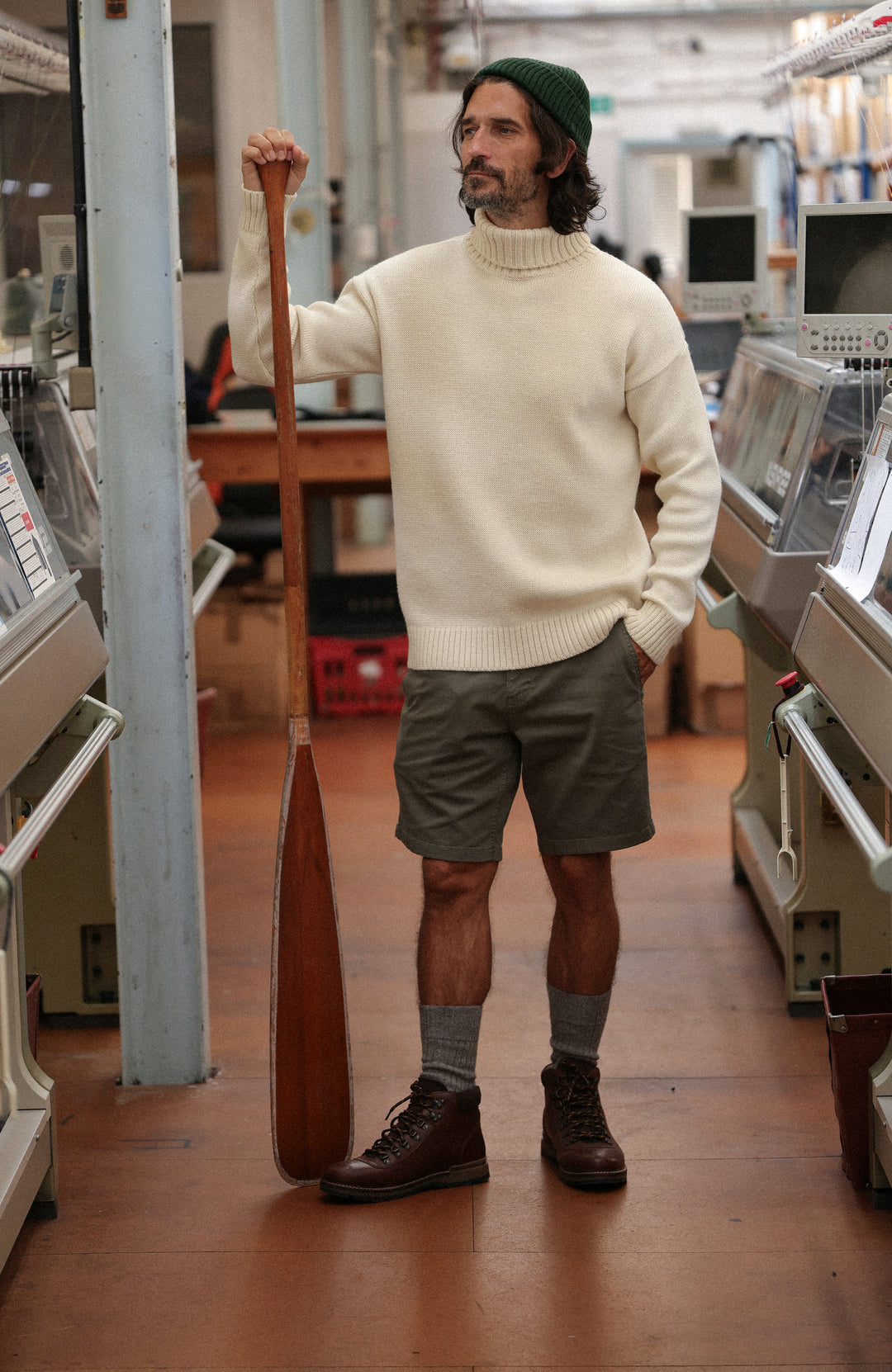 A man stands in a workshop holding a wooden paddle, wearing a cream-colored merino wool submariner jumper by Paul James Knitwear made in England turtleneck sweater, olive green shorts, a dark green beanie, gray socks, and brown boots.