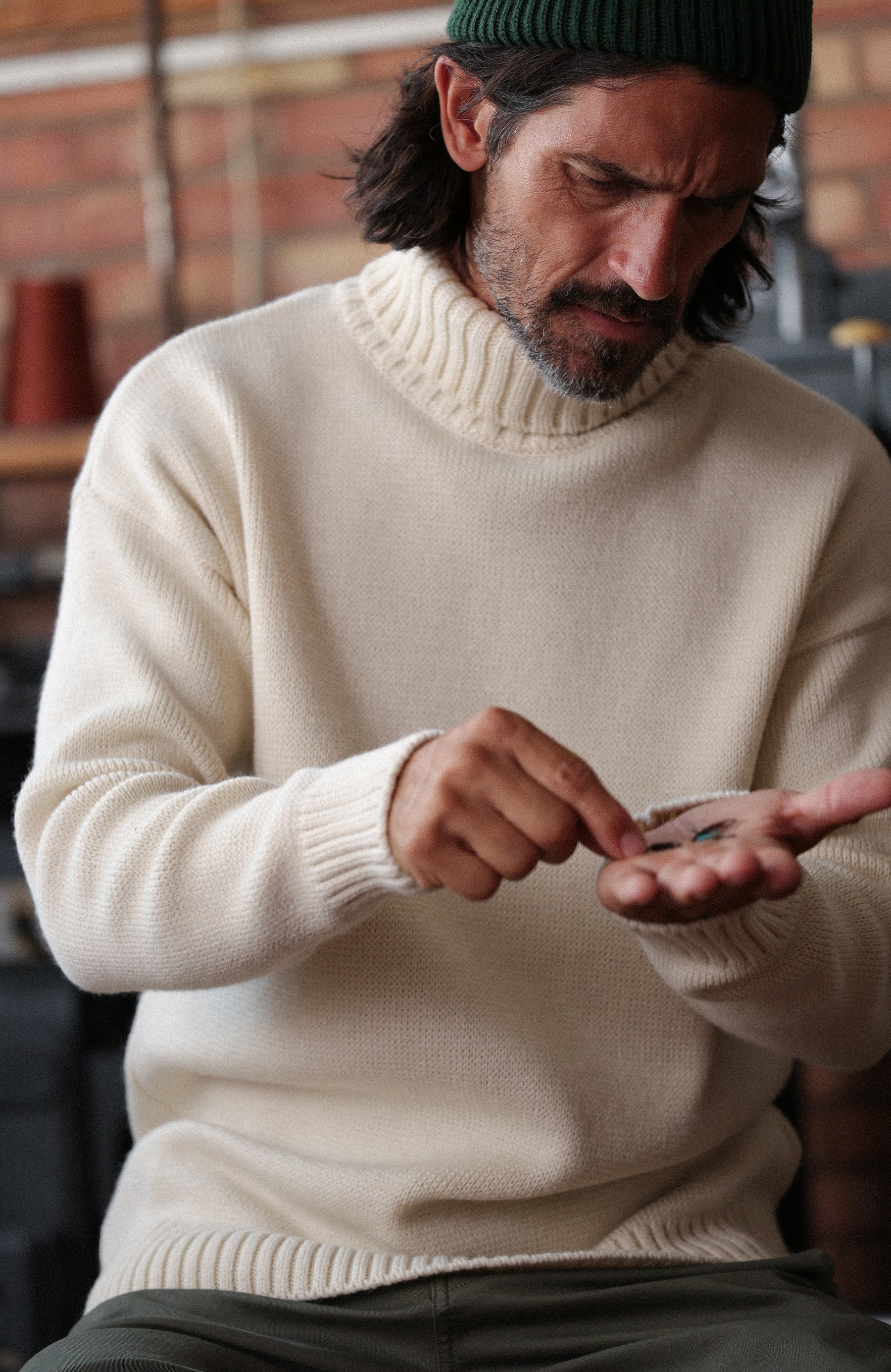 A man wearing a cream-colored merino wool submariner jumper by Paul James Knitwear made in England turtleneck sweater and a dark green beanie, looking down at his hands in a warm, rustic indoor setting.