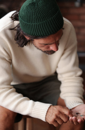 A man wearing a dark green ribbed knit fisherman wool beanie by Paul James Knitwear and a cream-colored knit sweater, looking down while holding something in his hands.