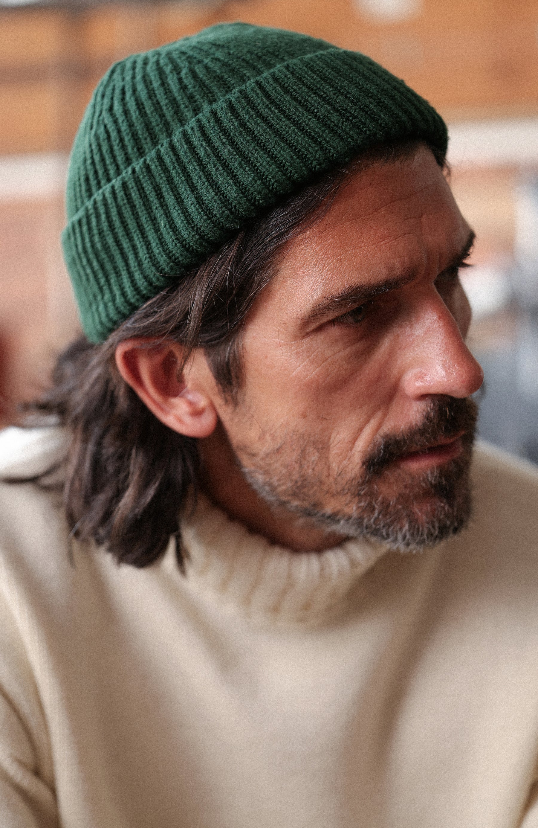 A man wearing a dark green merino wool fisherman beanie by Paul James Knitwear ribbed knit beanie and a cream-colored knit sweater, shown in a close-up profile view against a softly blurred indoor background