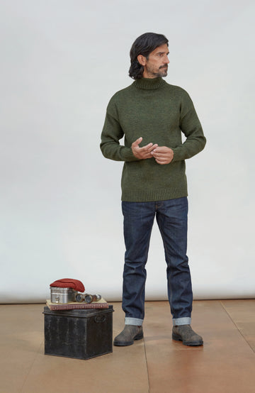 Made in England wool submariner by Paul James Knitwear.  A man wearing an olive green knit turtleneck sweater, dark blue jeans, and gray boots stands in a studio setting next to a small black storage trunk with a red object on top.