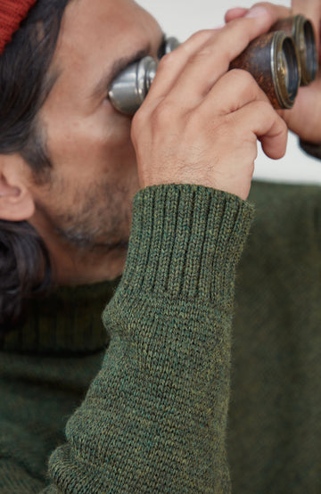Made in England wool submariner sweater by Paul James Knitwear. Close-up of a man wearing an olive green ribbed knit sweater and a red beanie, looking through vintage-style binoculars.