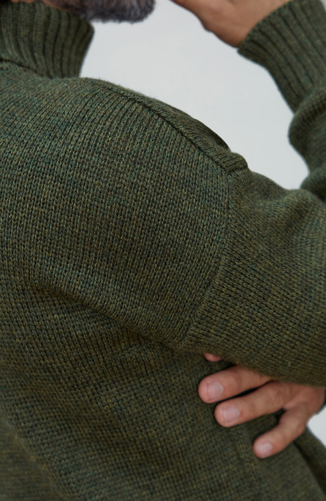 Made in England wool submariner sweater by Paul James Knitwear. Close-up of a person wearing a dark olive green ribbed knit sweater, showing the texture of the fabric and the sleeve cuff.