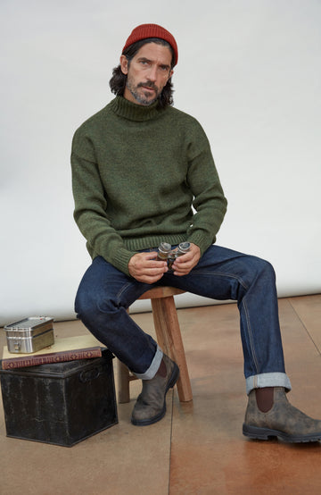 Made in England wool submariner sweater by Paul James Knitwear. A man sits on a wooden stool wearing an olive green knit turtleneck sweater, blue jeans, brown boots, and a red beanie in a studio setting.