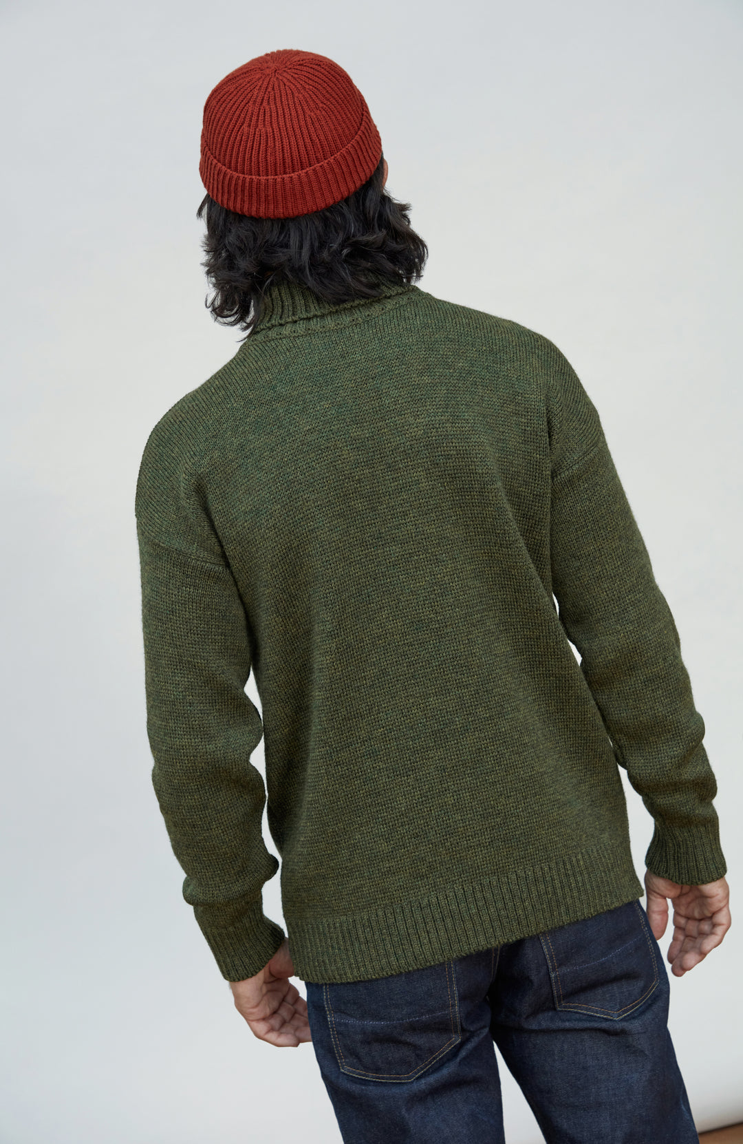 Made in England wool submariner sweater by Paul James Knitwear. A person shown from the back wearing an olive green knit sweater, a rust red beanie, and dark blue jeans against a plain light gray studio background.