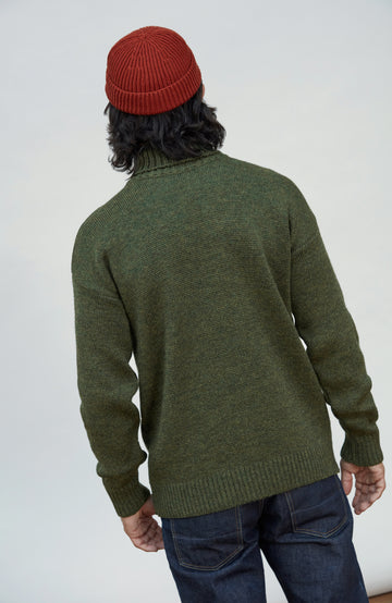 Made in England wool submariner sweater by Paul James Knitwear. A person shown from the back wearing an olive green knit sweater, a rust red beanie, and dark blue jeans against a plain light gray studio background.