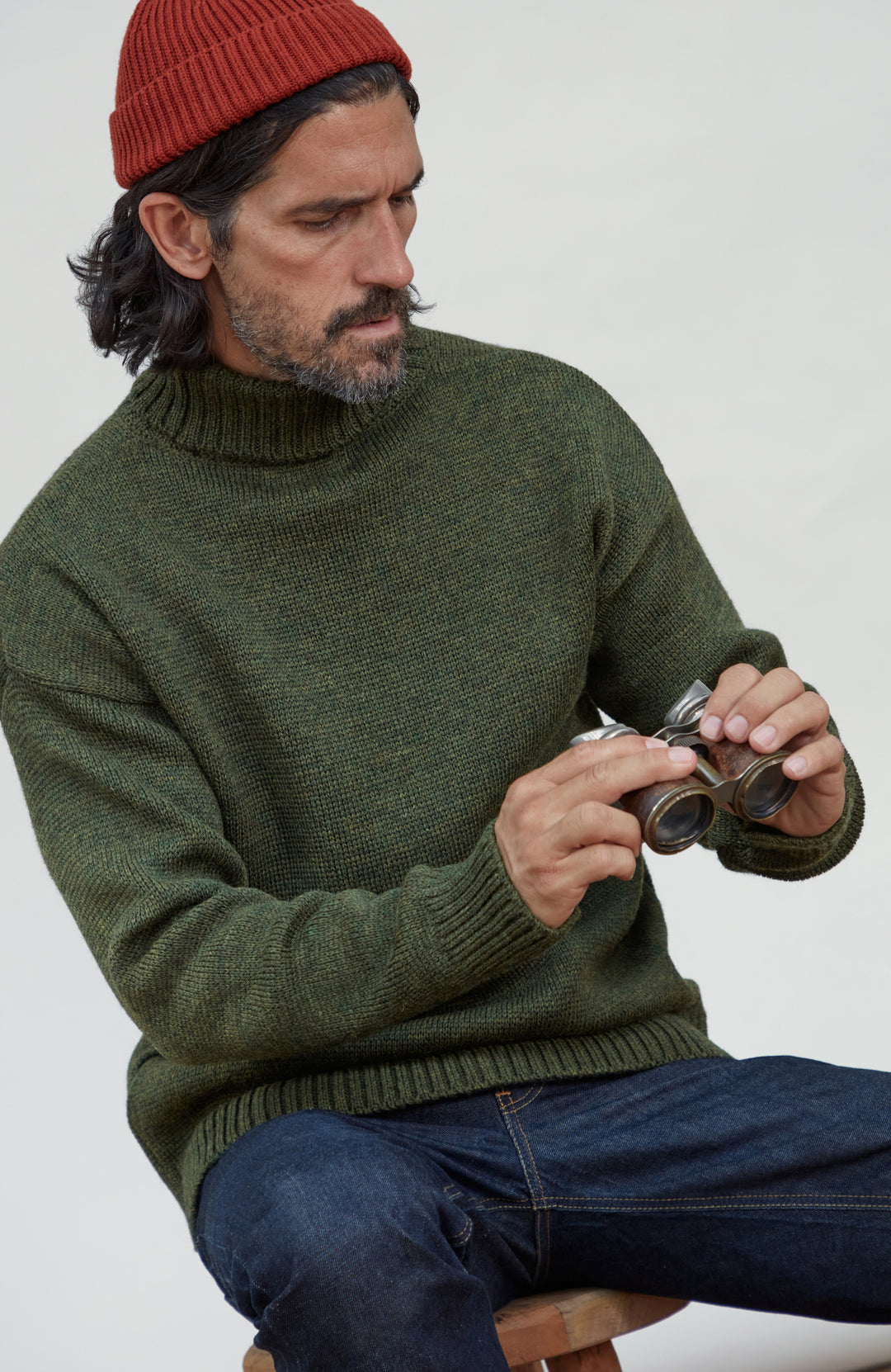 Made in England wool submariner by Paul James Knitwear. A man wearing an olive green knit turtleneck sweater, a red beanie, and blue jeans, holding a pair of vintage binoculars while seated against a plain light background.