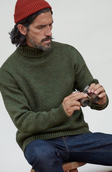 Made in England wool submariner by Paul James Knitwear. A man wearing an olive green knit turtleneck sweater, a red beanie, and blue jeans, holding a pair of vintage binoculars while seated against a plain light background.