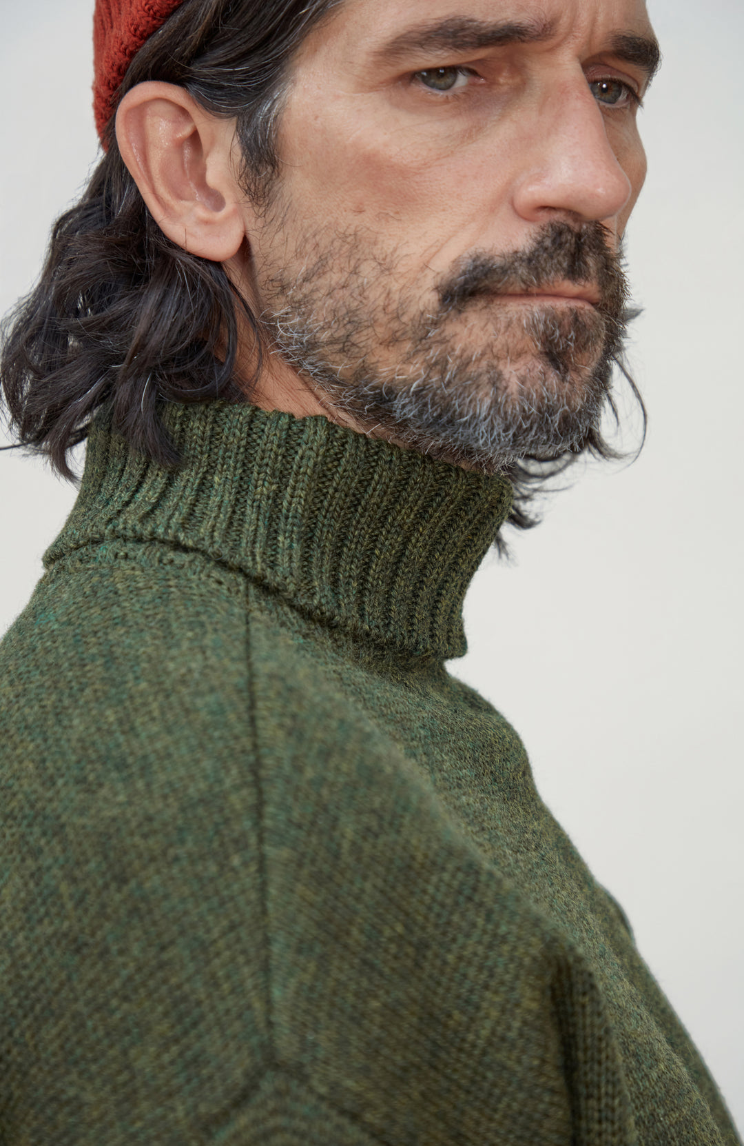 Made in England wool submariner sweater by Paul James Knitwear.  A close-up side profile of a man wearing a textured olive green ribbed turtleneck sweater and a rust-red knit beanie against a plain light background.