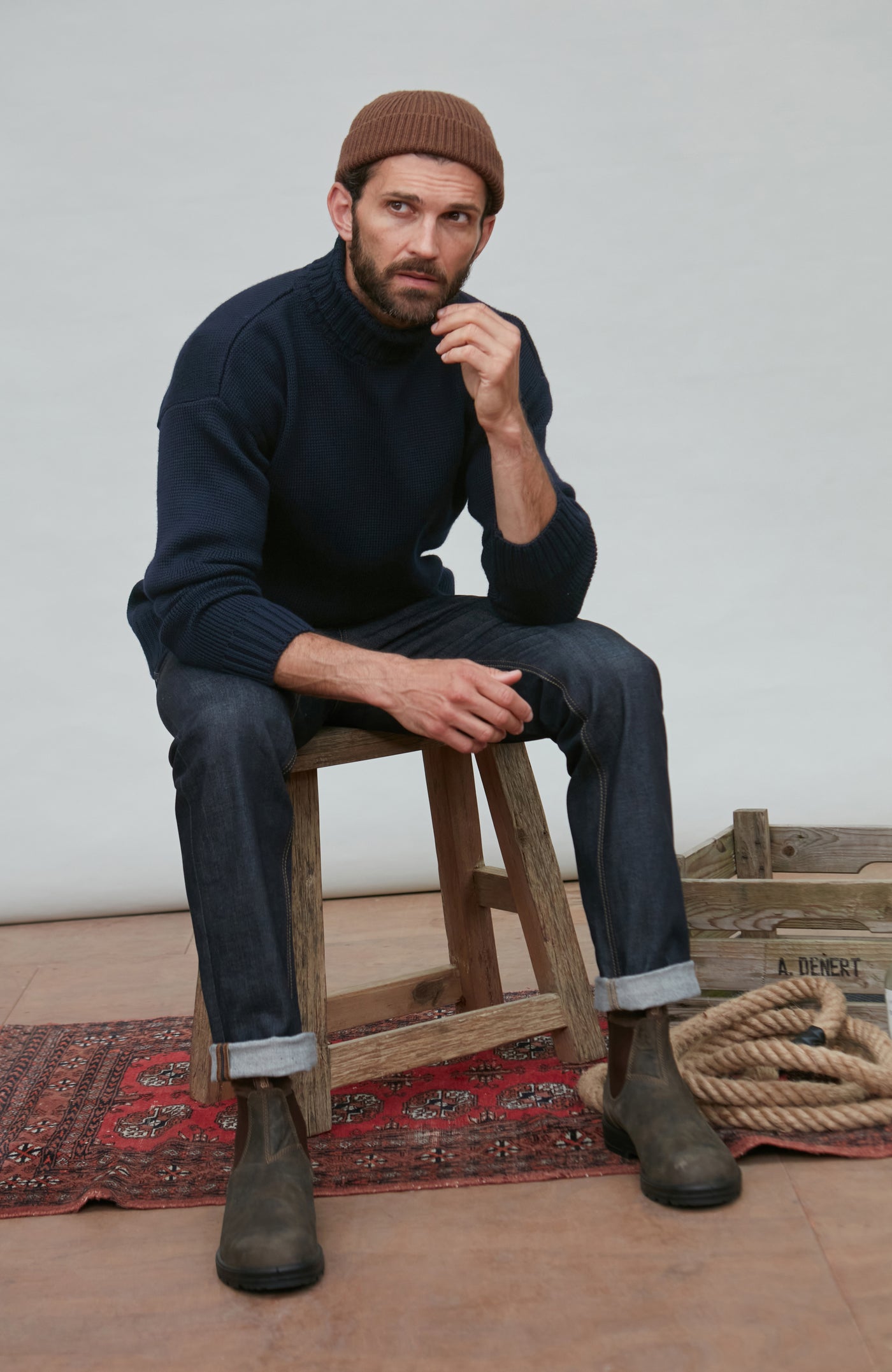 Man sitting on a wooden stool wearing a navy wool turtleneck sweater, brown beanie, and dark jeans.