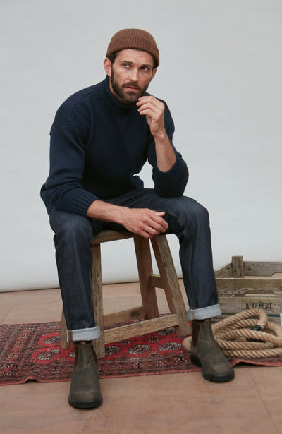 Man sitting on a wooden stool wearing a navy wool turtleneck sweater, brown beanie, and dark jeans.