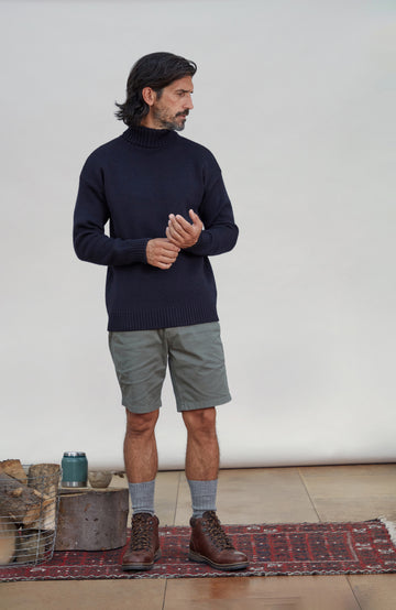 A man wearing a dark navy merino wool Submariner turtleneck sweater, olive green shorts, grey socks, and brown lace-up boots, standing on a patterned rug against a plain light grey studio background. Made by Paul James Knitwear.