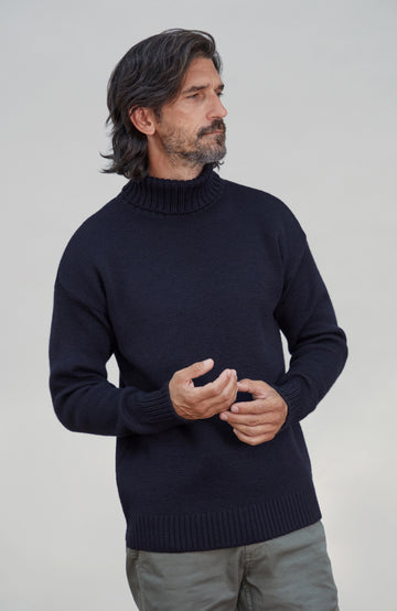 A man wearing a dark navy blue merino wool ribbed mock neck sweater, paired with gray trousers, standing against a light gray studio background. Made by Paul James Knitwear.