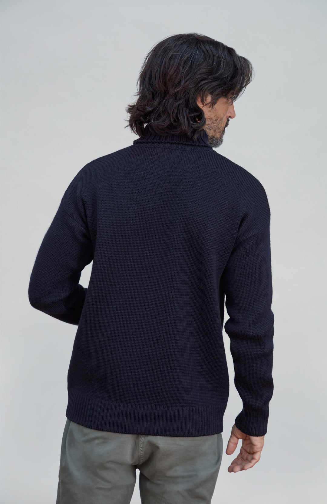 A man with dark hair is shown from the back wearing a dark navy blue knit sweater and gray trousers, photographed against a light gray studio background. a chunky wool sweater by Paul James Knitwear