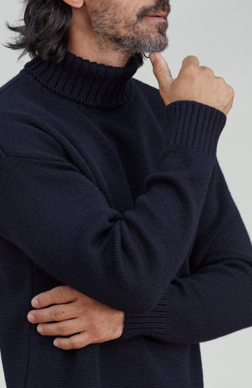 A close-up of a man wearing a dark navy blue ribbed knit turtleneck sweater, showing the texture of the fabric and the collar detail. Made by Paul James Knitwear.  Submariner sweater