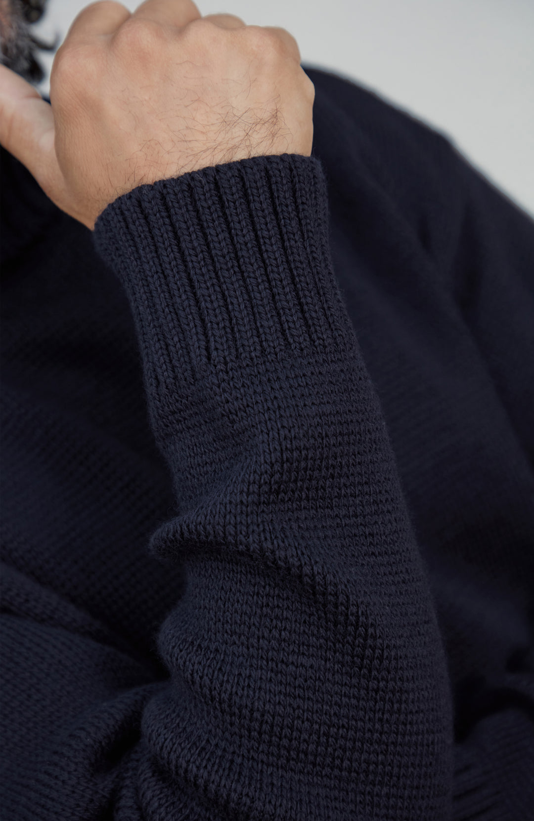 Close-up of a person's hand and forearm wearing a dark navy ribbed knit sweater, showing the texture of the knit fabric.Made by Paul James Knitwear. Submariner wool sweater