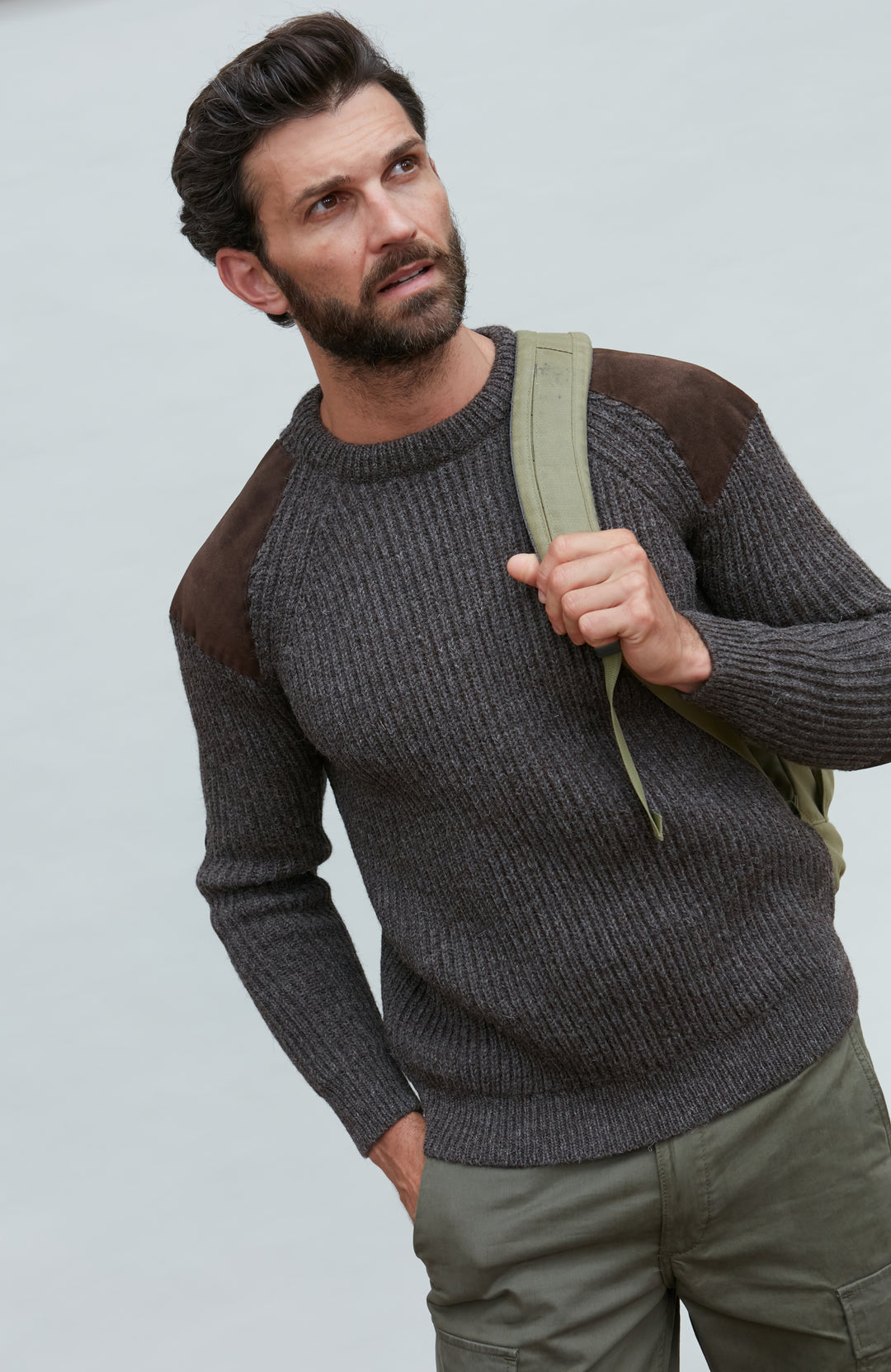 Man wearing a brown wool sweater with patches, holding a green backpack against a light gray background by Paul James
