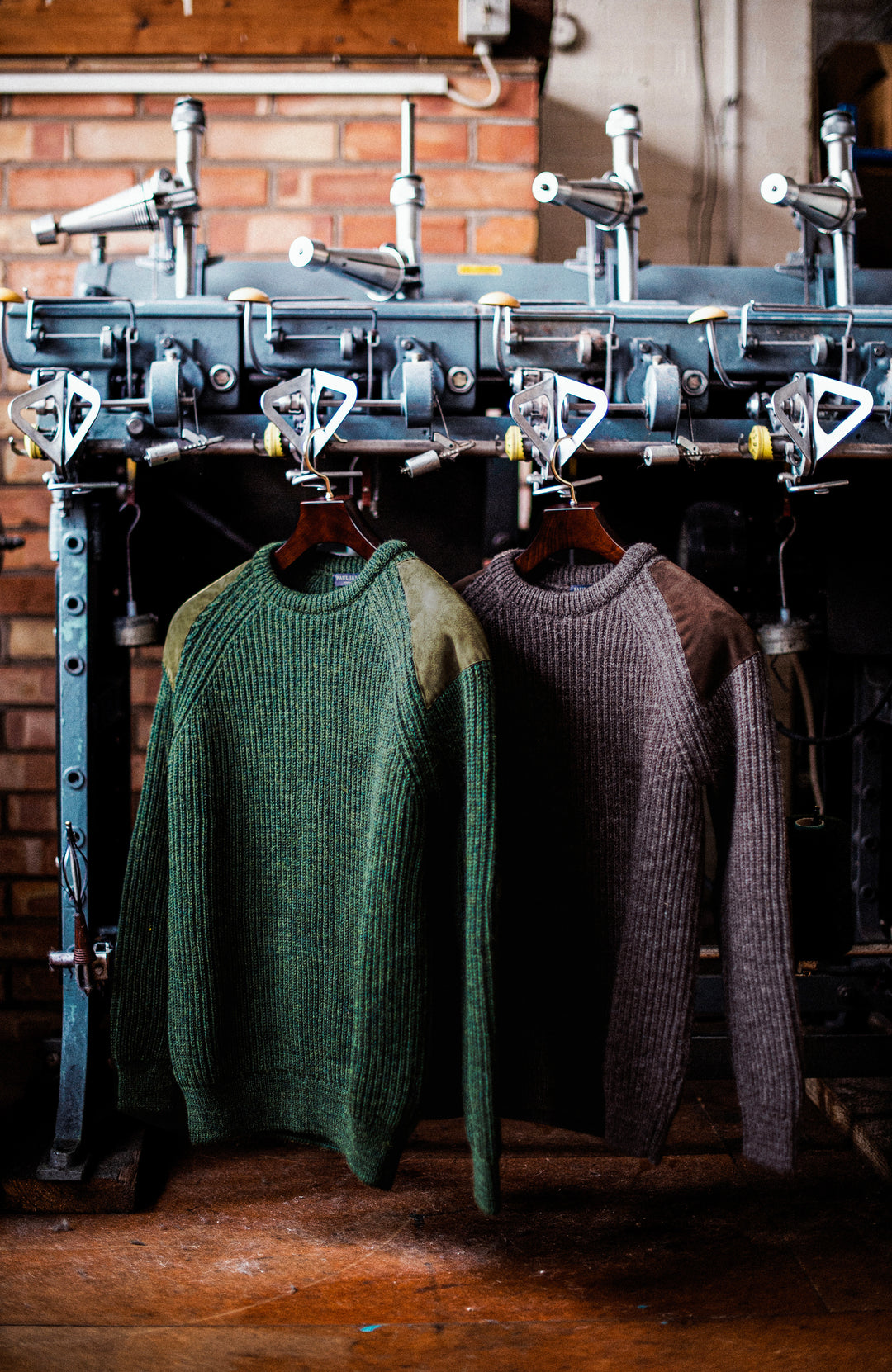 Two ribbed British wool shooting knit crewneck sweaters by Paul James Knitwear hang on hangers in front of a vintage industrial garment press. The sweater on the left is forest green with tan shoulder panels, and the sweater on the right is a dark taupe-brown.