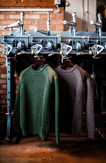 Two ribbed British wool shooting knit crewneck sweaters by Paul James Knitwear hang on hangers in front of a vintage industrial garment press. The sweater on the left is forest green with tan shoulder panels, and the sweater on the right is a dark taupe-brown.