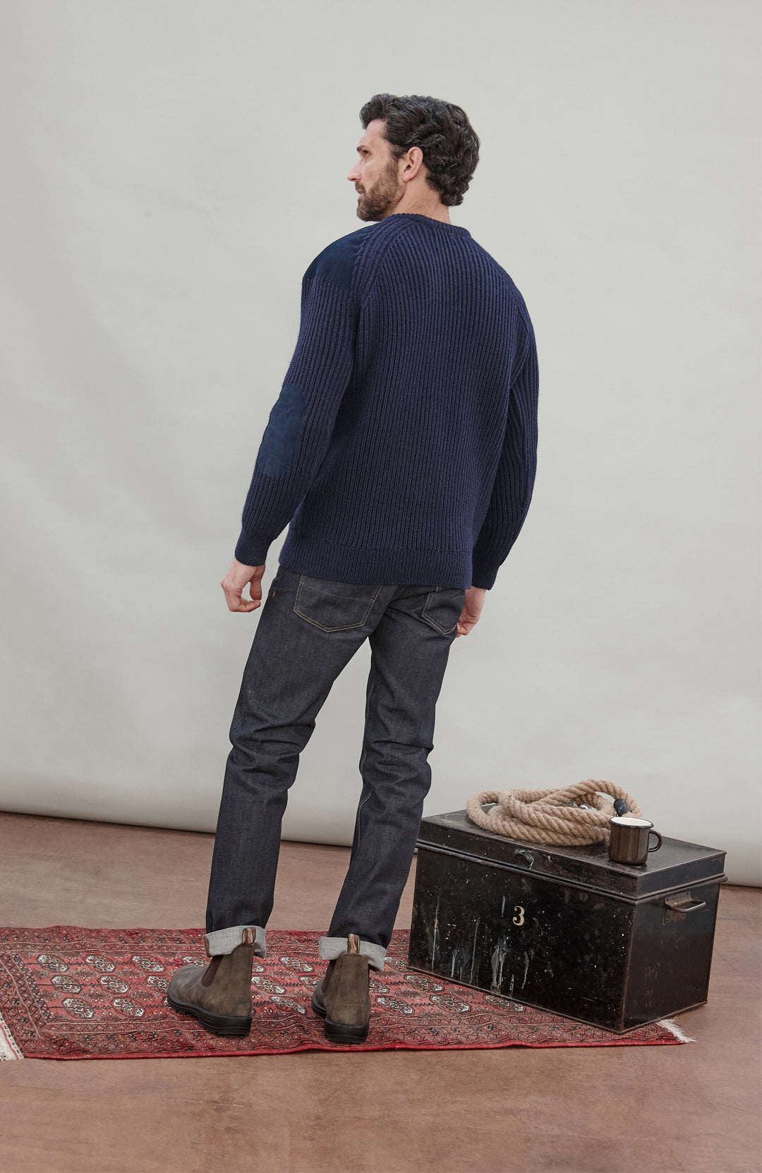 Man wearing a navy sweater and dark pants standing next to a black trunk with a rope on a red rug.