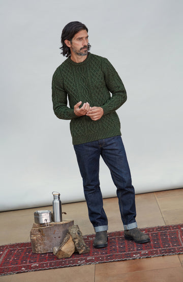 British wool sweater made in England by Paul James Knitwear. A man wearing a dark green cable-knit sweater, dark blue jeans, and dark boots stands on a patterned rug in front of a light gray studio backdrop. A small metal container and wood block sit on the floor to his left.