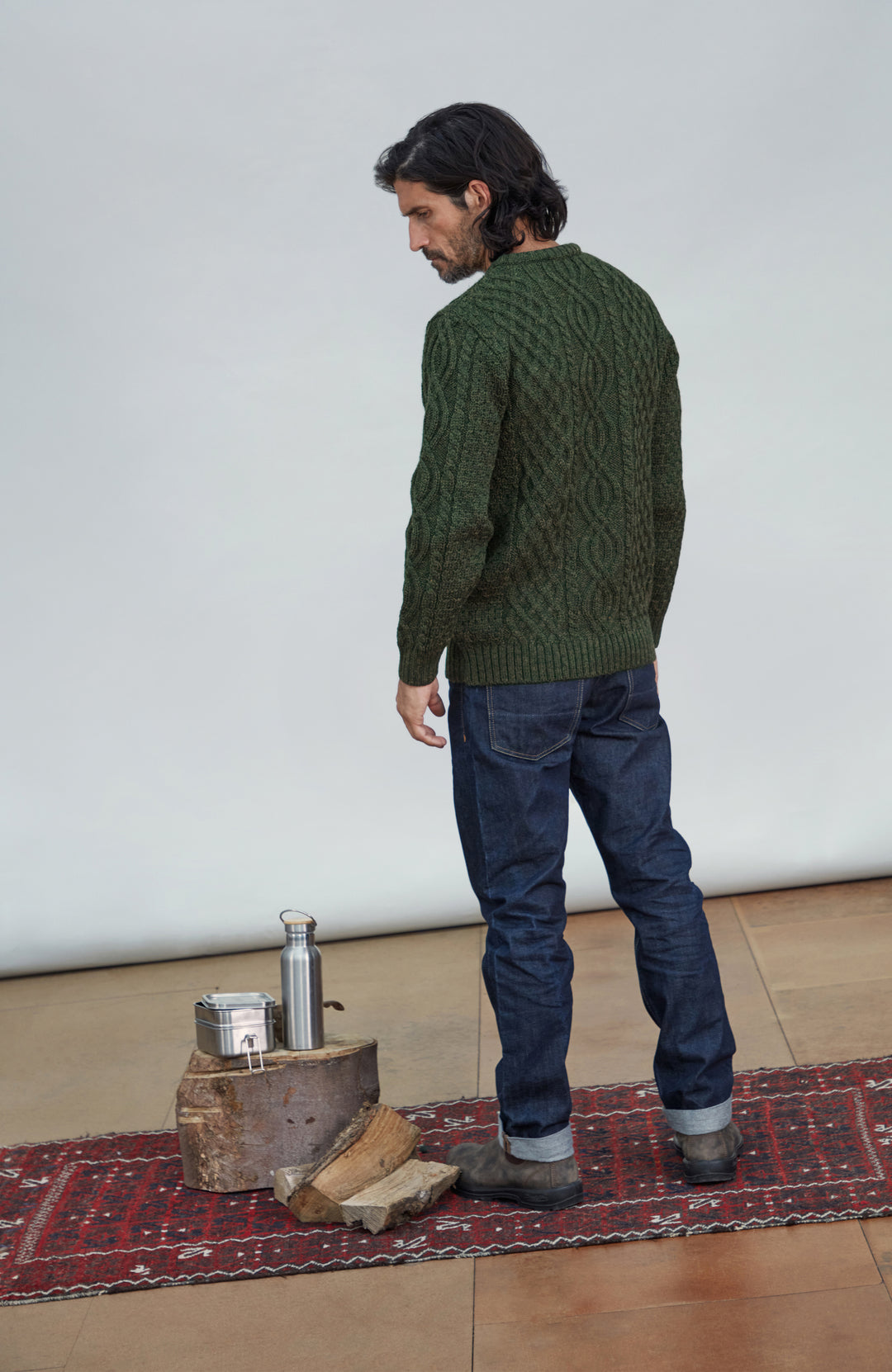 British wool sweater made in England by Paul James Knitwear. A man wearing a dark green cable-knit sweater and blue jeans stands on a red patterned rug in a studio setting, looking down. A small wooden stand with a metal flask and box is beside him.