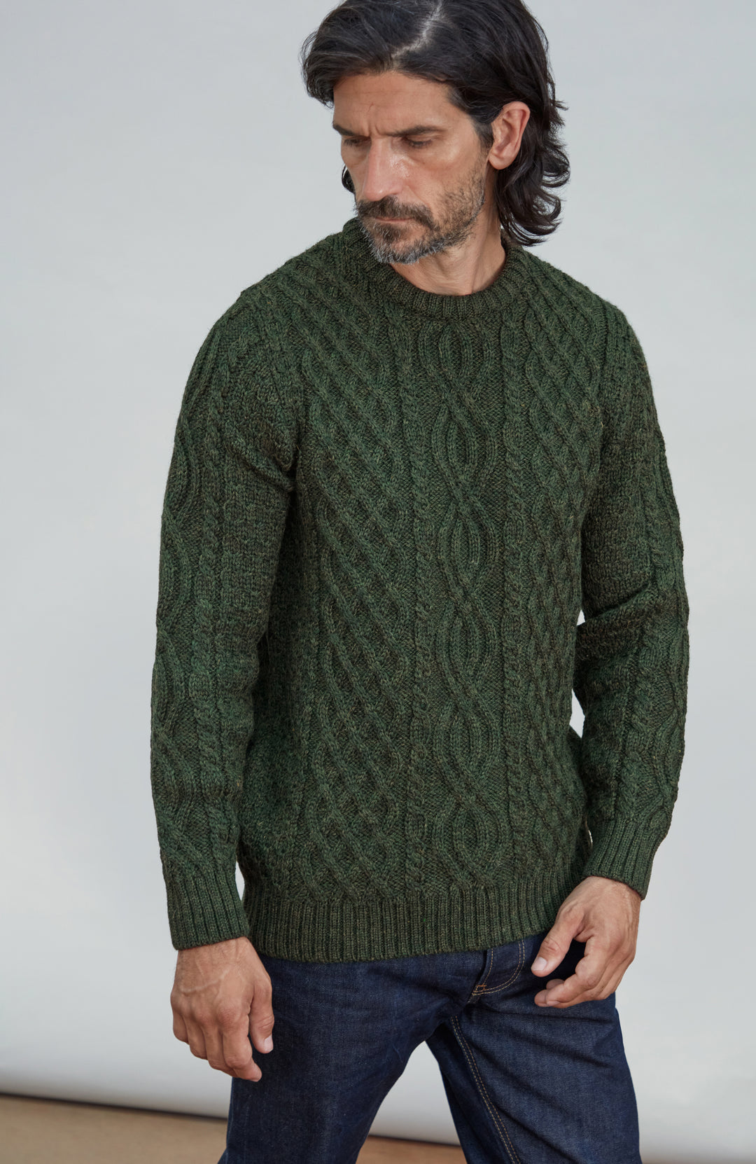 A man wearing a dark green made in England, British wool cable-knit sweater and dark blue jeans, standing against a plain light gray background. Made by Paul James Knitwear.