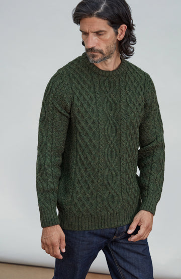 A man wearing a dark green made in England, British wool cable-knit sweater and dark blue jeans, standing against a plain light gray background. Made by Paul James Knitwear.