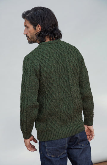 British wool sweater made in England by Paul James Knitwear. A man wearing a dark green cable-knit sweater and blue jeans, photographed from the back against a plain light gray studio background.