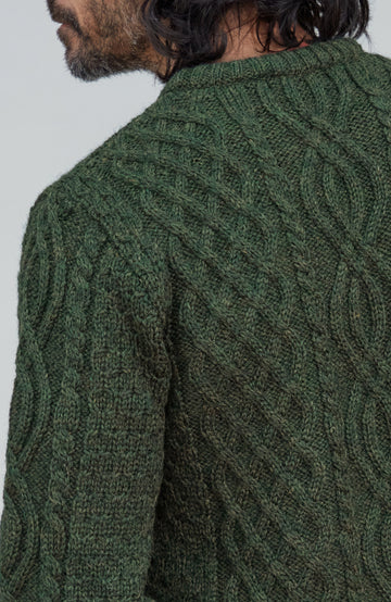 British wool sweater made in England by Paul James Knitwear. Close-up of a man wearing a dark green cable-knit sweater, showing the textured knit pattern and collar against a plain light gray background.