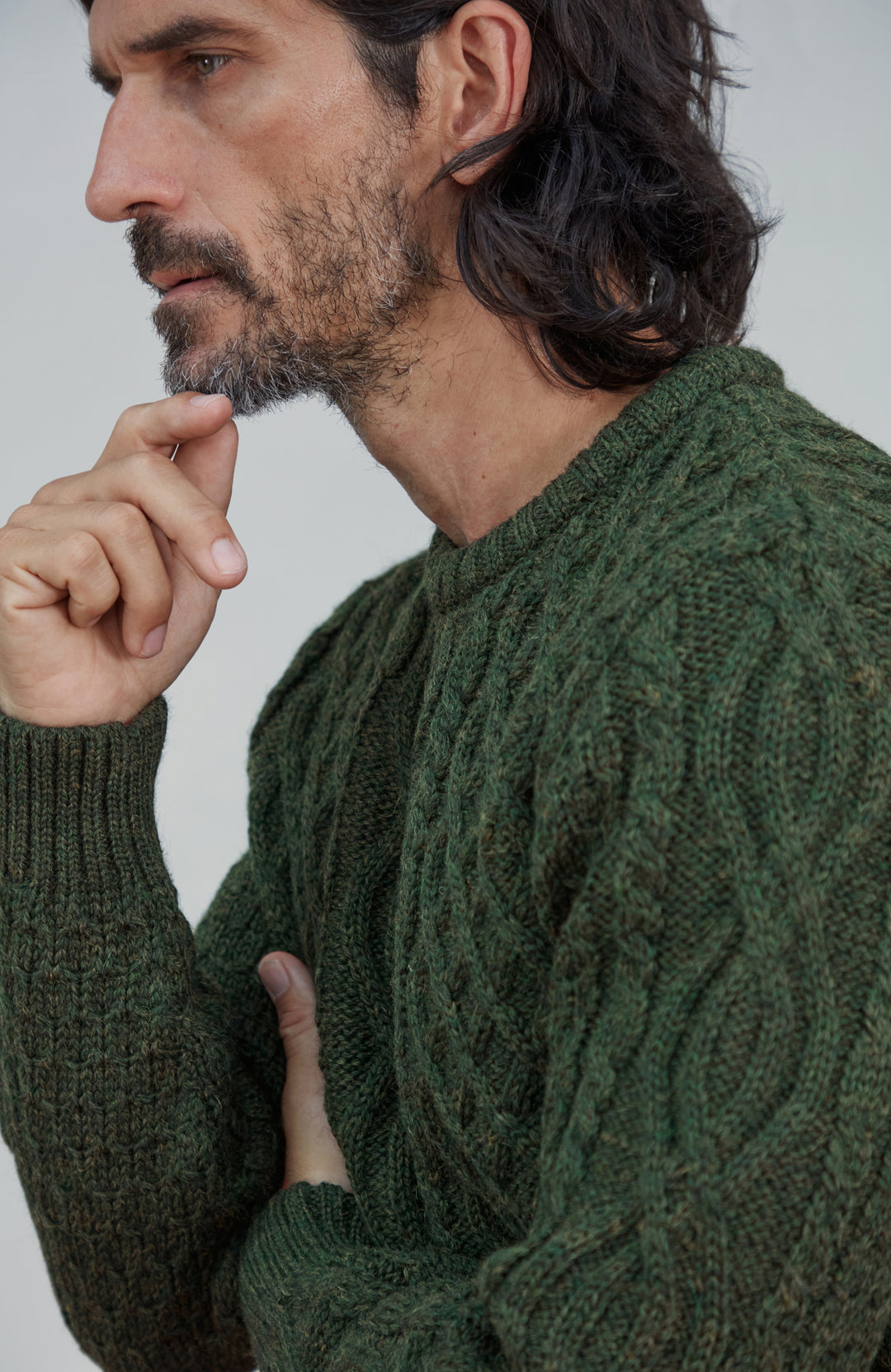 British wool sweater made in England by Paul James Knitwear. A close-up side profile of a man wearing a dark forest green cable-knit sweater against a light gray background.