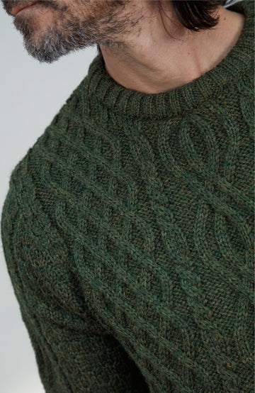 British wool sweater made in England by Paul James Knitwear. Close-up of a man wearing a dark olive green cable-knit sweater, showing the detailed knit pattern and collar against a light gray background.