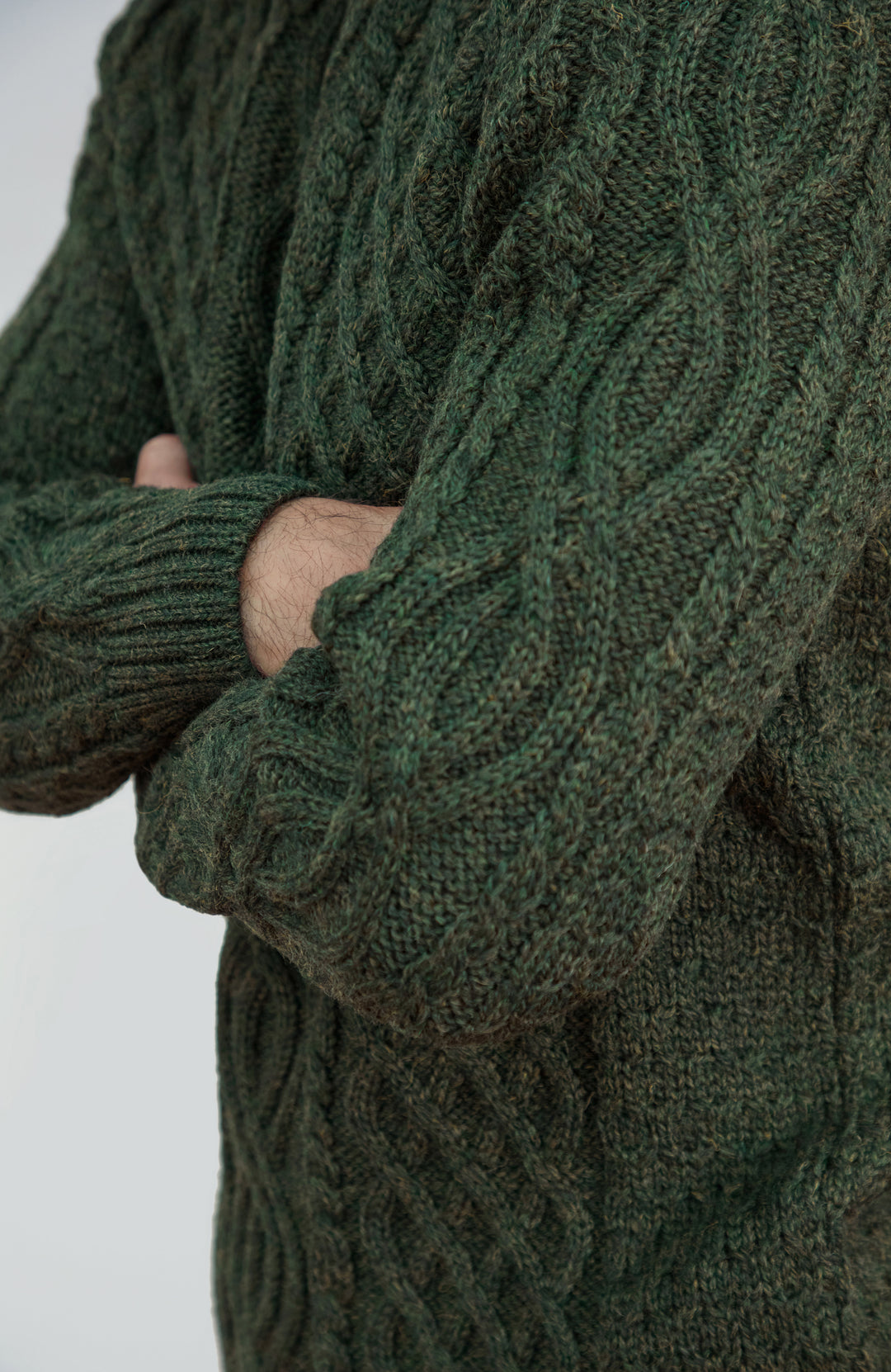 British wool sweater made in England by Paul James Knitwear. Close-up of a person wearing a dark forest green cable-knit sweater, showing the detailed knit texture and crossed arms against a light gray background.