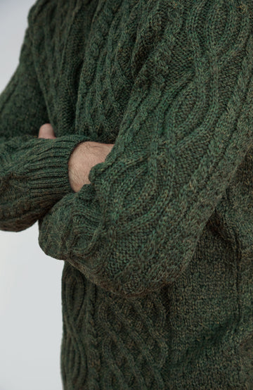 British wool sweater made in England by Paul James Knitwear. Close-up of a person wearing a dark forest green cable-knit sweater, showing the detailed knit texture and crossed arms against a light gray background.