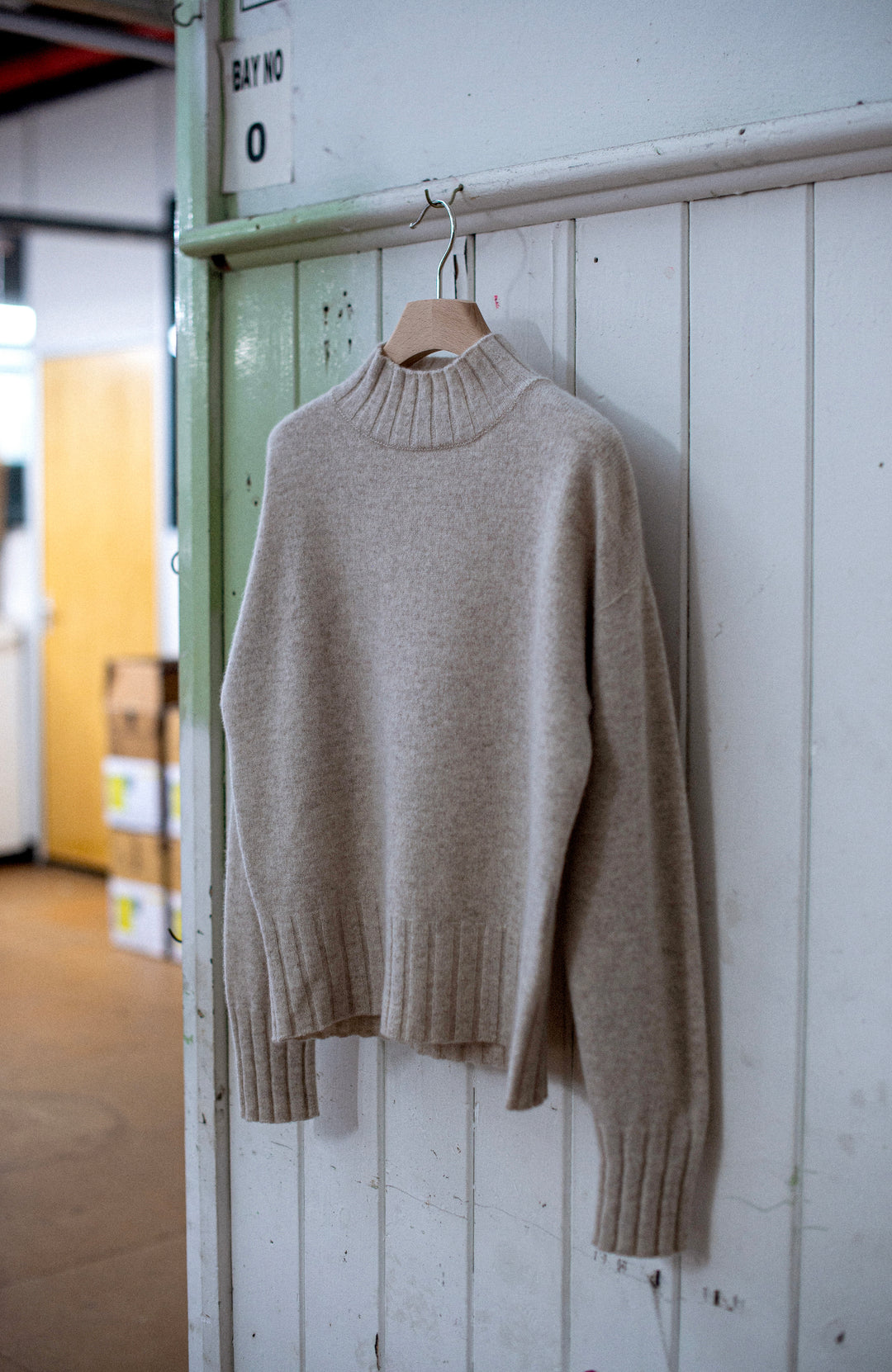 A light beige womens knit mock-neck  wool sweater hanging on a wooden hanger against a white paneled wall in an indoor industrial setting. Made in England by Paul James Knitwear. 