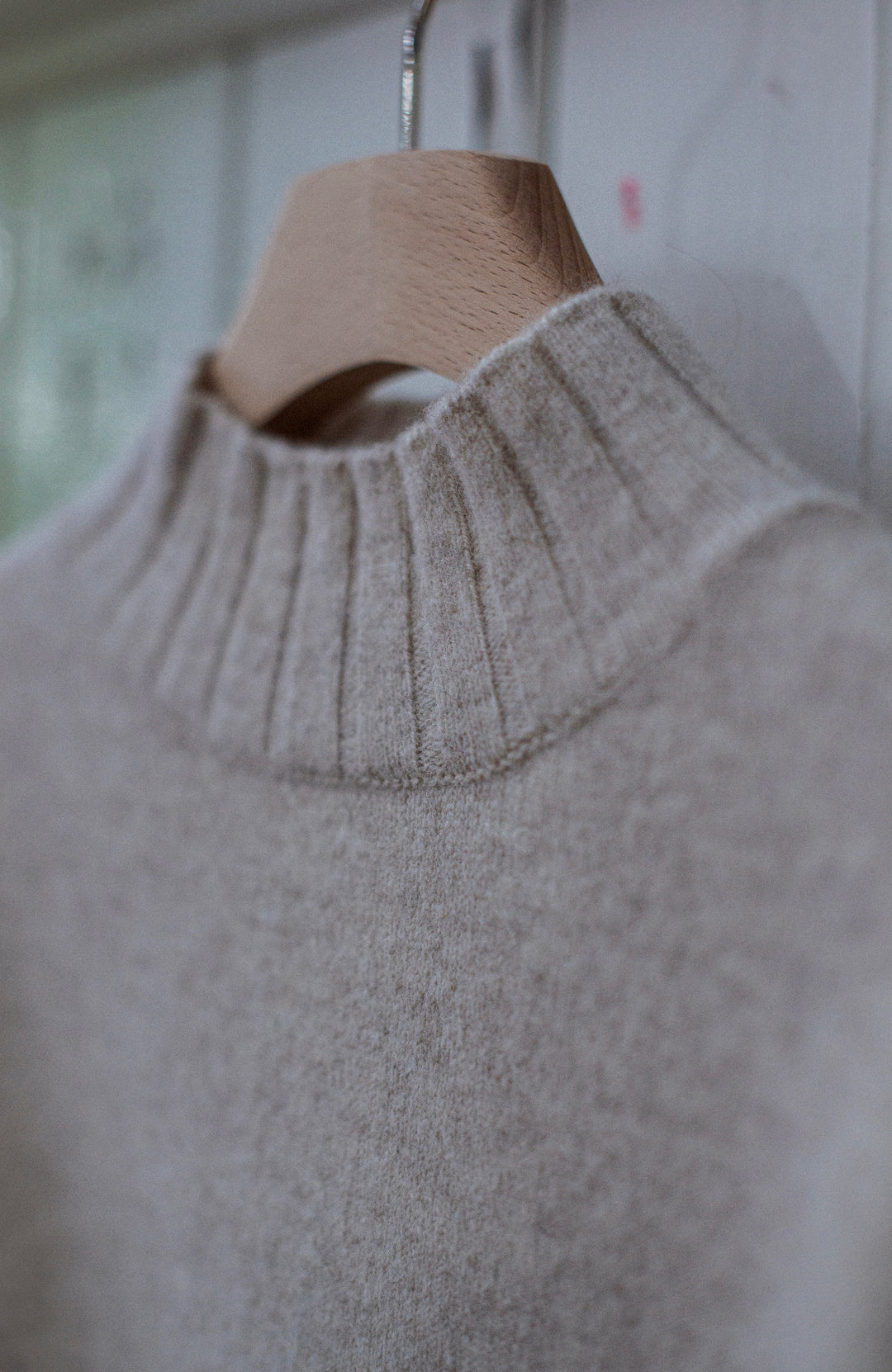 A close-up of a light beige knit sweater with a ribbed mock neck, hanging on a wooden hanger against a soft-focus indoor background. Jumper made in England by Paul James Knitwear, lambswool