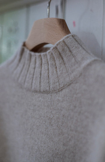 A close-up of a light beige knit sweater with a ribbed mock neck, hanging on a wooden hanger against a soft-focus indoor background. Jumper made in England by Paul James Knitwear, lambswool