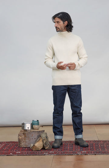 Men's Made in England Chunky Merino Wool Submariner Jumper .A man wearing a cream ribbed turtleneck sweater, dark blue jeans, and dark brown boots stands on a patterned rug in front of a light gray wall. A rustic wooden crate with a metal canister and a small green ceramic cup sits on the floor to his left.