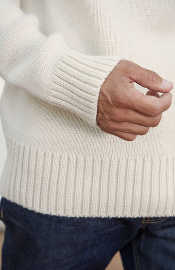 Men's Made in England Chunky Merino Wool Submariner Jumper .Close-up of a cream-colored ribbed knit sweater worn by a person, showing the sleeve cuff, hand, and hem, paired with dark blue jeans.