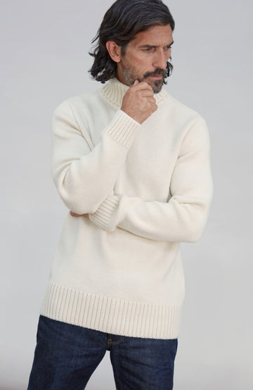 Men's Made in England Chunky Merino Wool Submariner Jumper .A man wearing a cream-colored ribbed mock neck sweater and dark blue jeans, standing against a light gray studio background.