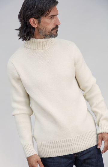 Men's Made in England Chunky Merino Wool Submariner Jumper . A man wearing a cream-colored ribbed mock neck sweater, paired with dark blue jeans, photographed against a neutral light gray background.