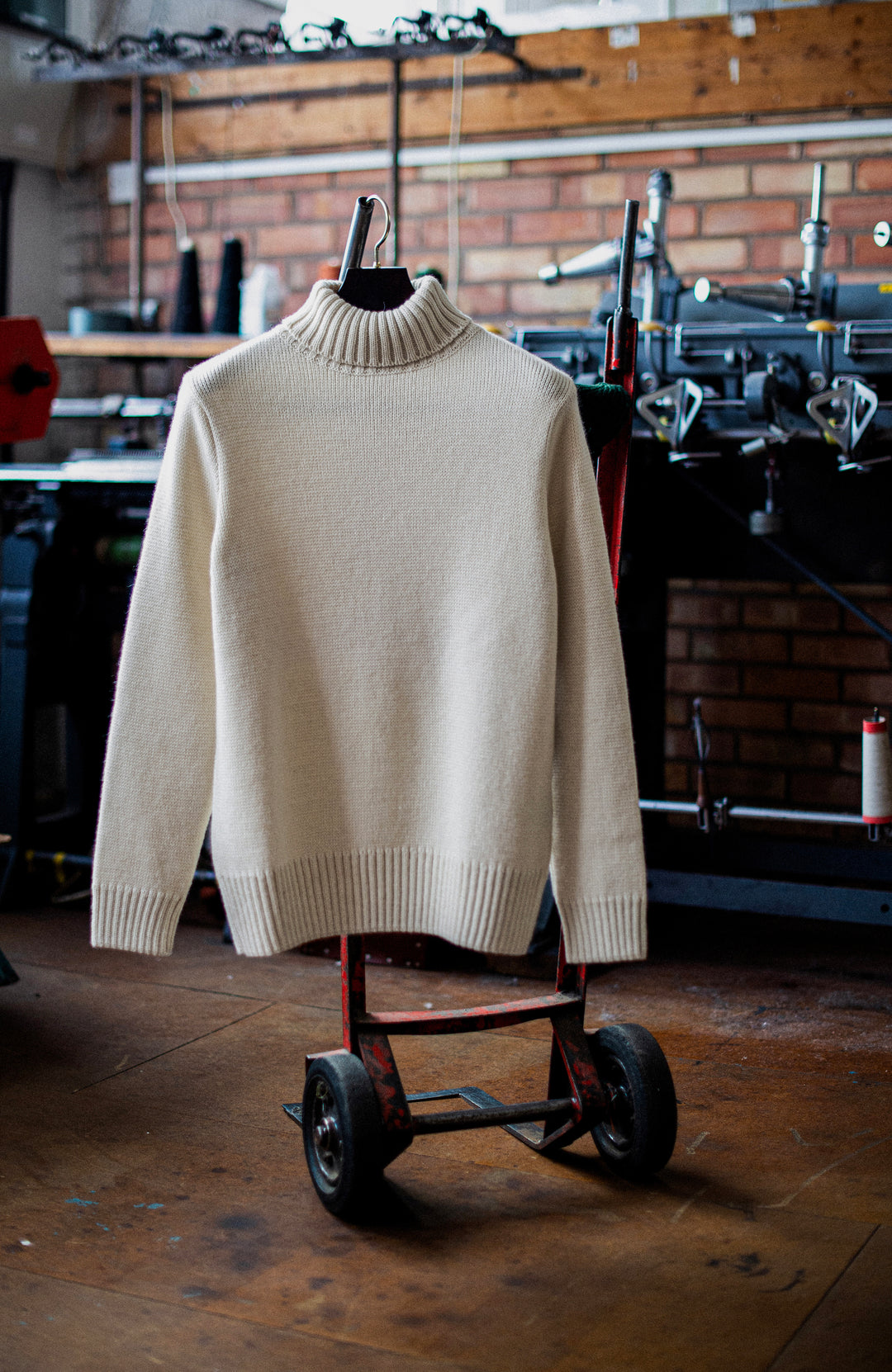 A cream-colored knit mock neck sweater hanging on a black hanger from a red metal cart in a workshop setting with a brick wall and tools in the background. Made in England wool sweater by Paul James Knitwear