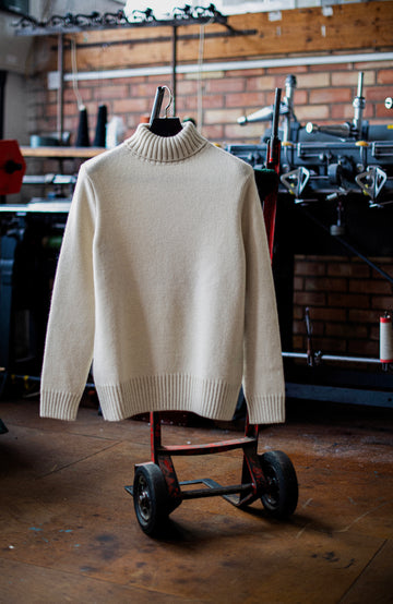 A cream-colored knit mock neck sweater hanging on a black hanger from a red metal cart in a workshop setting with a brick wall and tools in the background. Made in England wool sweater by Paul James Knitwear