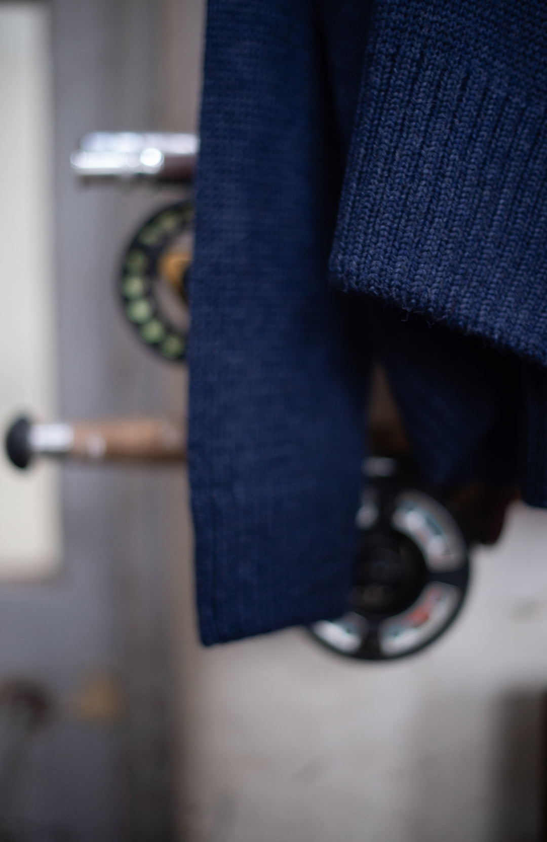 Blue wool made in england roll neck sweater by paul james knitwear hanging on a wooden coat rack with fishing equipment in the background
