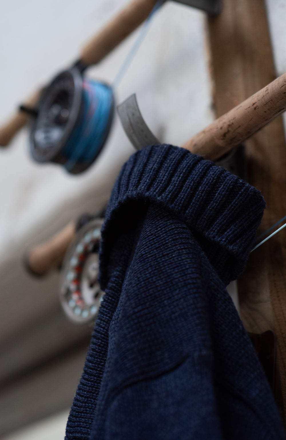 Blue wool made in england roll neck sweater by paul james knitwear hanging on a wooden coat rack with fishing equipment in the background