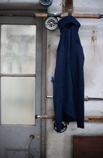 Blue wool made in england roll neck sweater by paul james knitwear hanging on a wooden coat rack with fishing equipment in the background