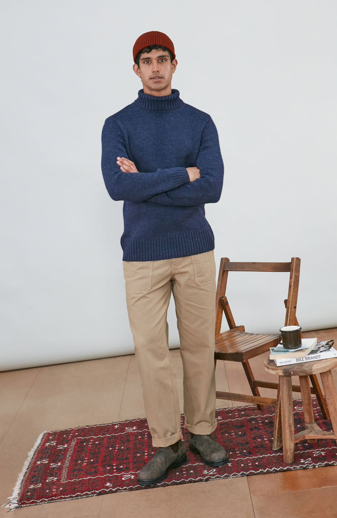 A man wearing a navy blue knit wool turtleneck sweater made in england by paul james knitwear, a rust-orange ribbed merino wool beanie, and beige trousers, posed against a light gray studio background.