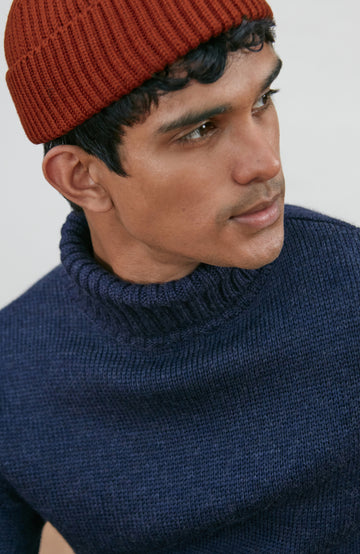 A man wearing a rust-orange ribbed knit fine merino wool beanie by paul james knitwear and a navy blue turtleneck sweater, photographed against a light gray studio background.
