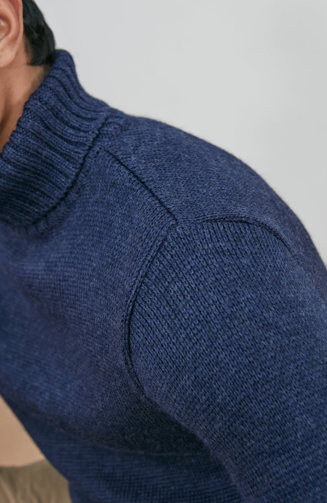 A man wearing a navy blue knit wool turtleneck sweater made in england by paul james knitwear, a rust-orange ribbed merino wool beanie, and beige trousers, posed against a light gray studio background.